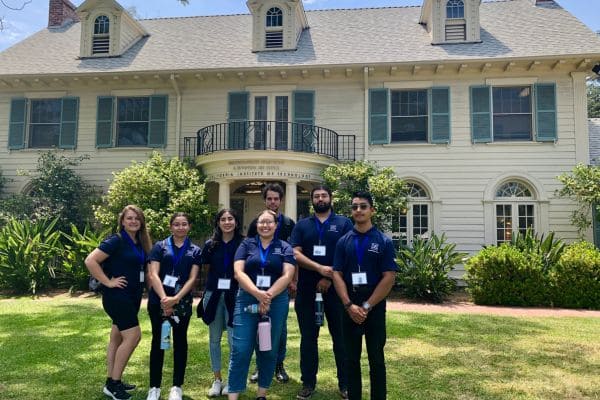 : A group of seven people in matching navy shirts stand smiling in front of a large, elegant, two-story building with a manicured lawn and green shutters.