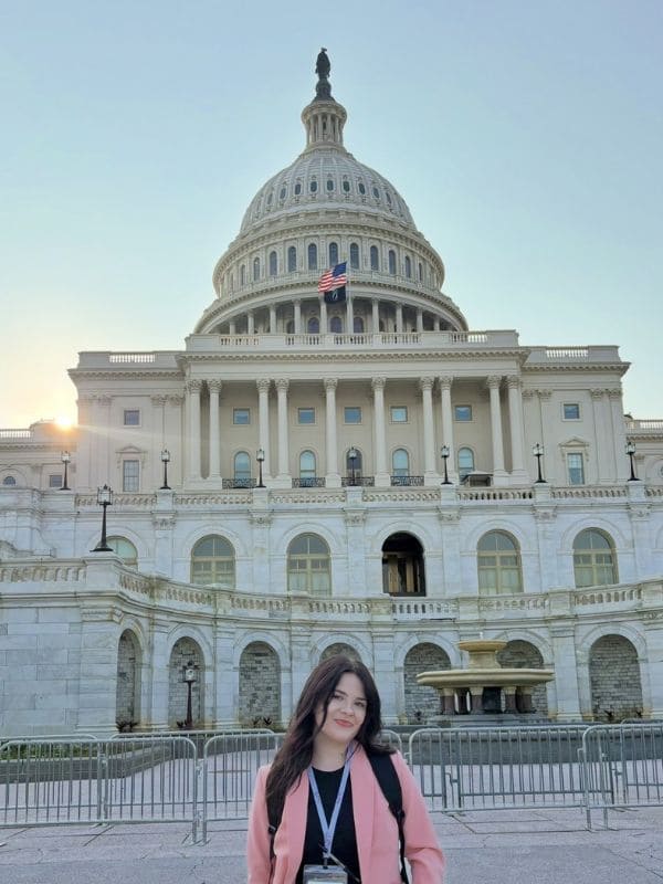 A woman stands smiling in front of the U.S. Capitol building, wearing a pink blazer and black top. The sun sets behind the iconic domed structure.