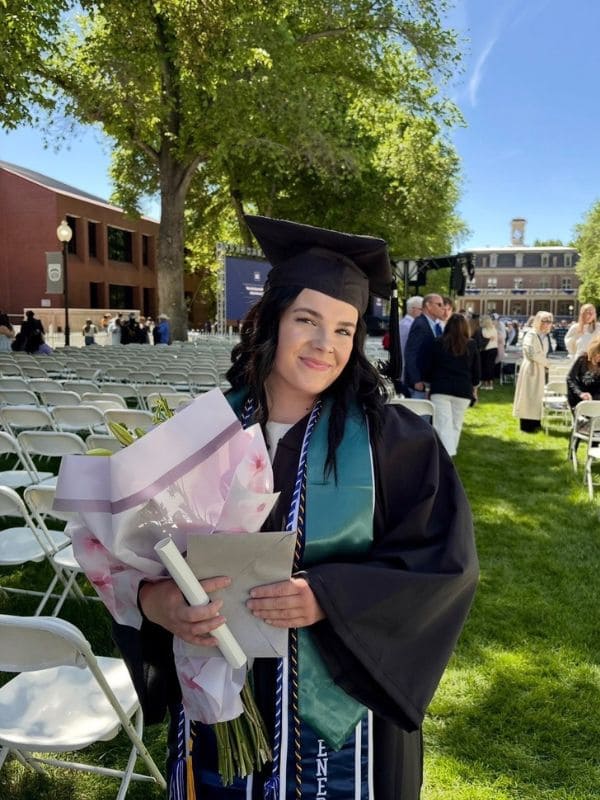 A graduate smiles, holding a diploma and flowers, wearing a cap and gown. She stands outdoors in a sunny ceremony, with white chairs and people in the background.