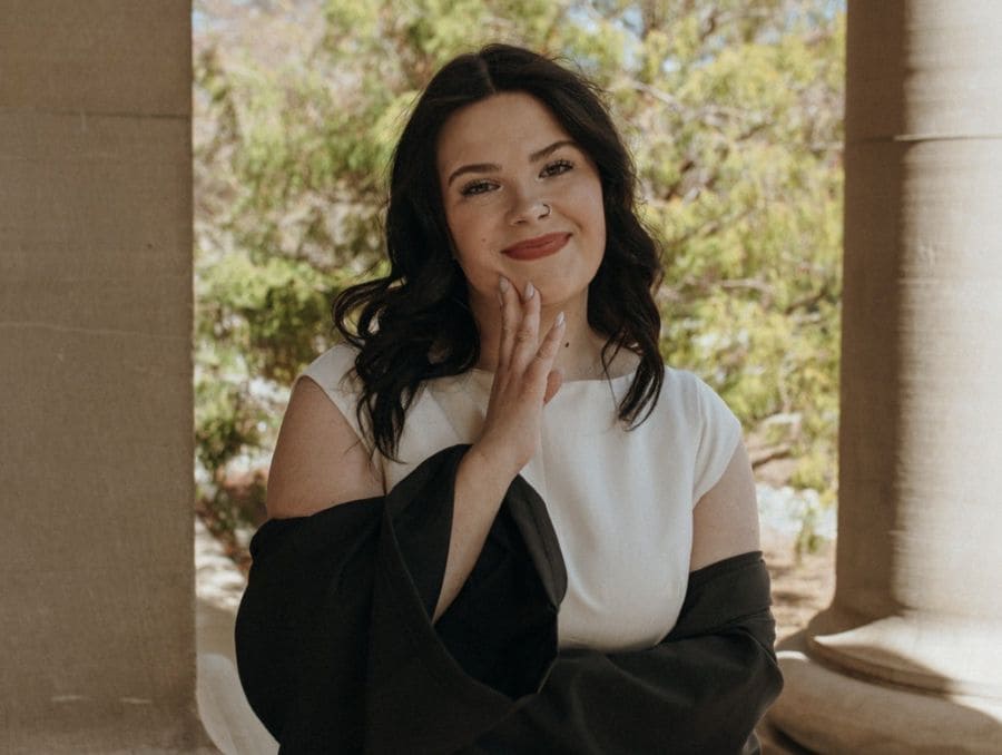Smiling woman with dark hair and white dress, draped in a black graduation gown. She’s standing in a columned outdoor setting.