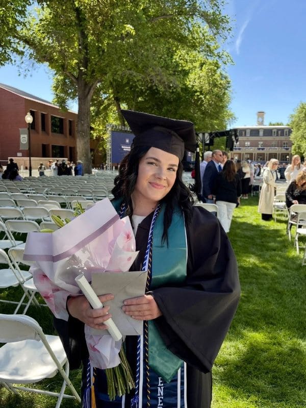 Fia Ewers wears a gown and holds a bouquet and a diploma, smiling outdoors on a sunny day. Rows of white chairs and people gathered in the background convey celebration.