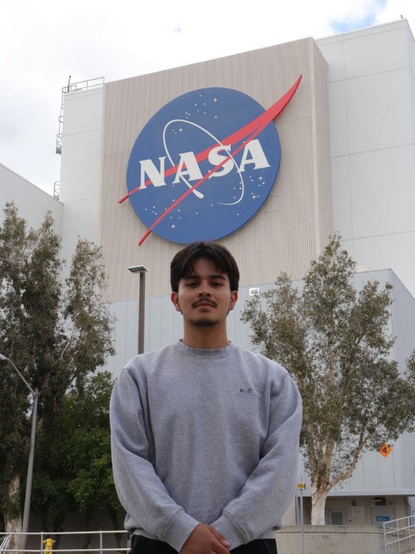 A person stands confidently in front of a building with a large NASA logo, surrounded by trees under a cloudy sky.