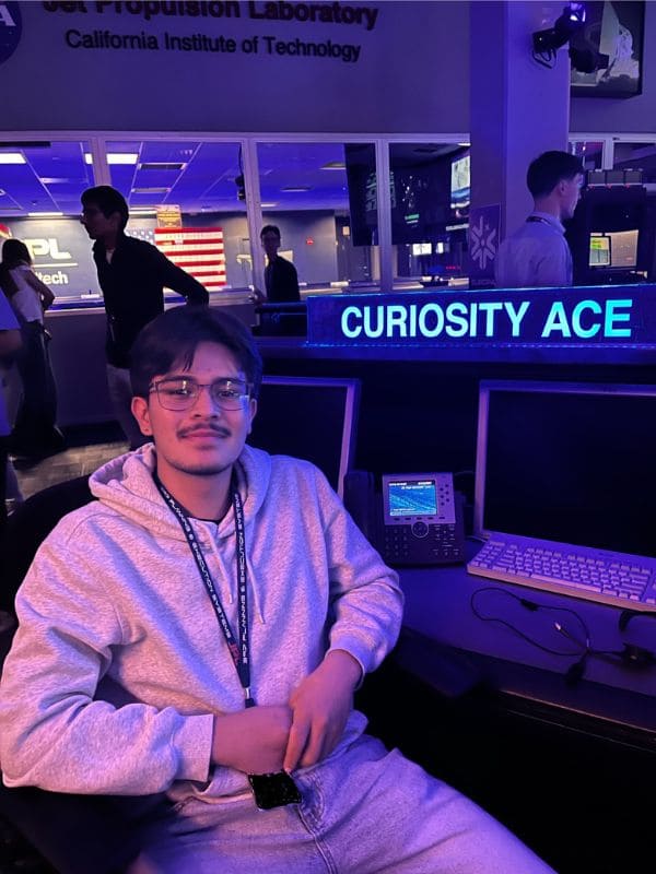 A person in glasses and a hoodie sits in a dimly lit control room at the Jet Propulsion Laboratory, with "Curiosity Ace" glowing on a nearby screen.