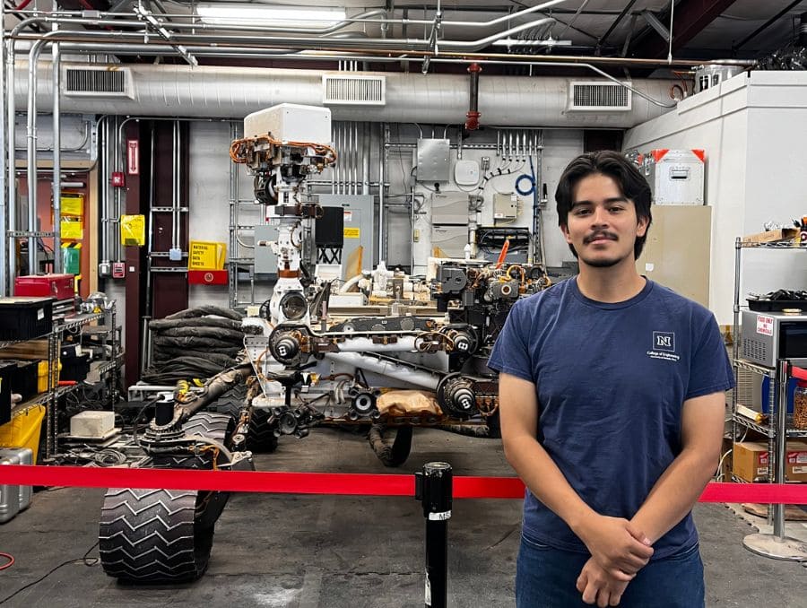 A man in a blue t-shirt stands in front of a Mars rover model inside a workshop filled with machinery and equipment, conveying a sense of innovation and exploration.