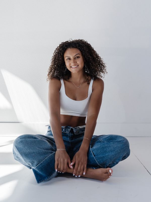 Jasmine Harris sits cross-legged on the floor in a sunlit room, wearing a white tank top and blue jeans, exuding a relaxed and cheerful vibe.