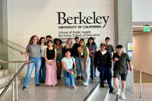 A diverse group of young adults stands on stairs, smiling, against a wall with "Berkeley University of California" text. Casual attire, bright atmosphere.
