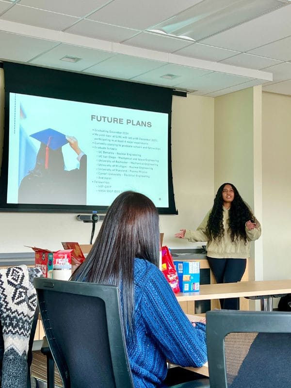 A woman presents in a classroom, gesturing toward a screen displaying "Future Plans." Attendees are seated at tables with snacks in the foreground.