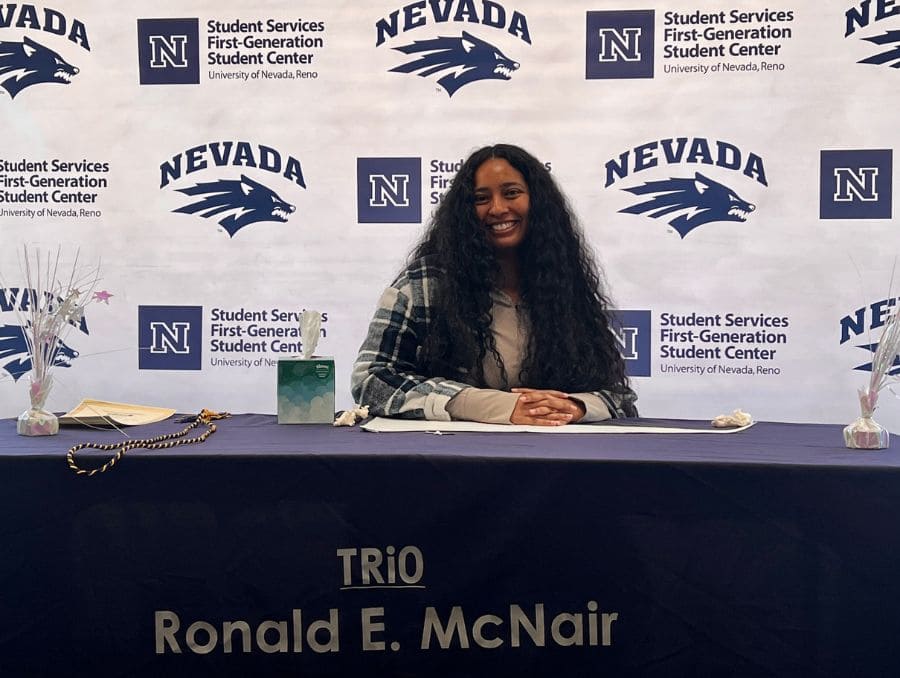 A woman with long hair smiles while seated at a table with a "TRiO Ronald E. McNair" banner. Behind her is a University of Nevada, Reno backdrop.