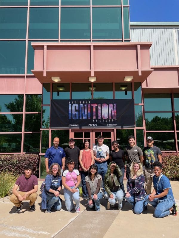 A group of fourteen people poses outside a building beneath a banner reading "National Ignition Facility" with Lawrence Livermore National Laboratory and NASA logos.