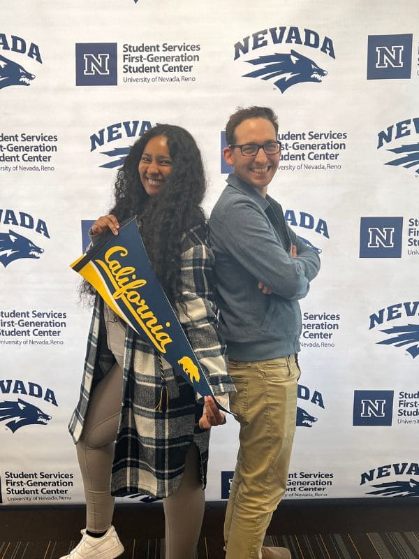 Two people stand back to back, smiling, in front of a University of Nevada, Reno backdrop. One holds a pennant that says "California.