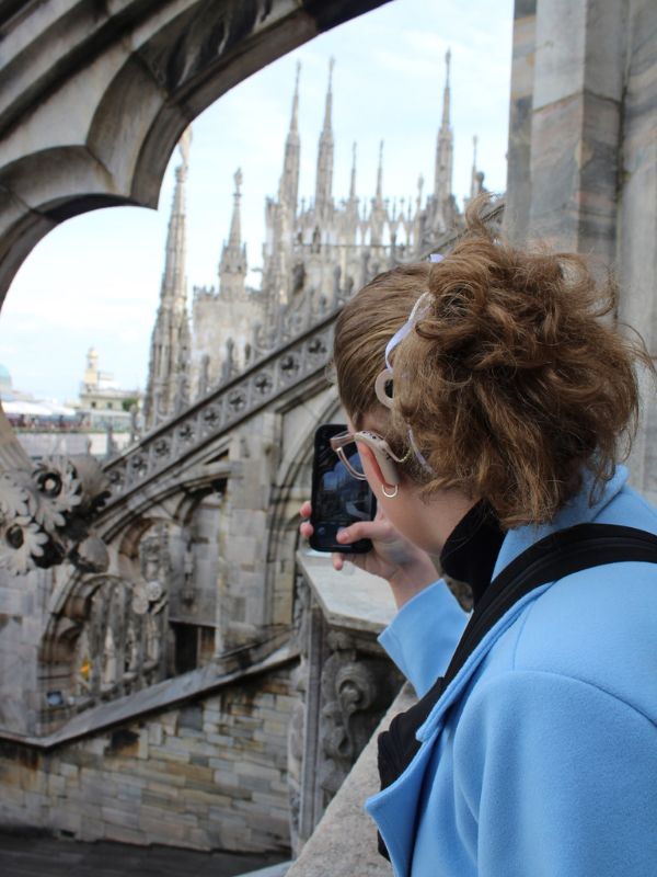 A person in a blue coat takes a photo with a smartphone, overlooking the intricate spires and arches of a Gothic cathedral.