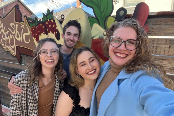 Four people smiling in front of a colorful mural featuring fruits. The sky is clear, and the mood is cheerful and friendly.