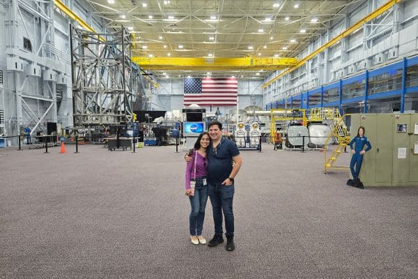 A smiling couple stands in a spacious facility with industrial equipment and a large American flag on the wall. The environment appears clean and well-lit.