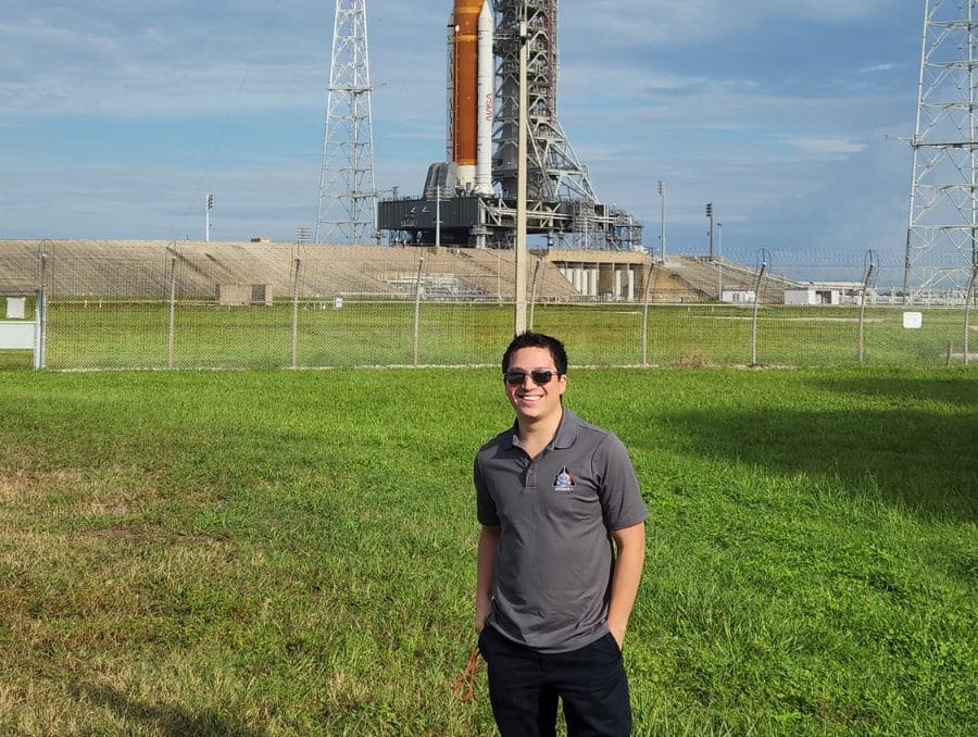 Manuel smiles in front of a space launch pad under a clear sky. 