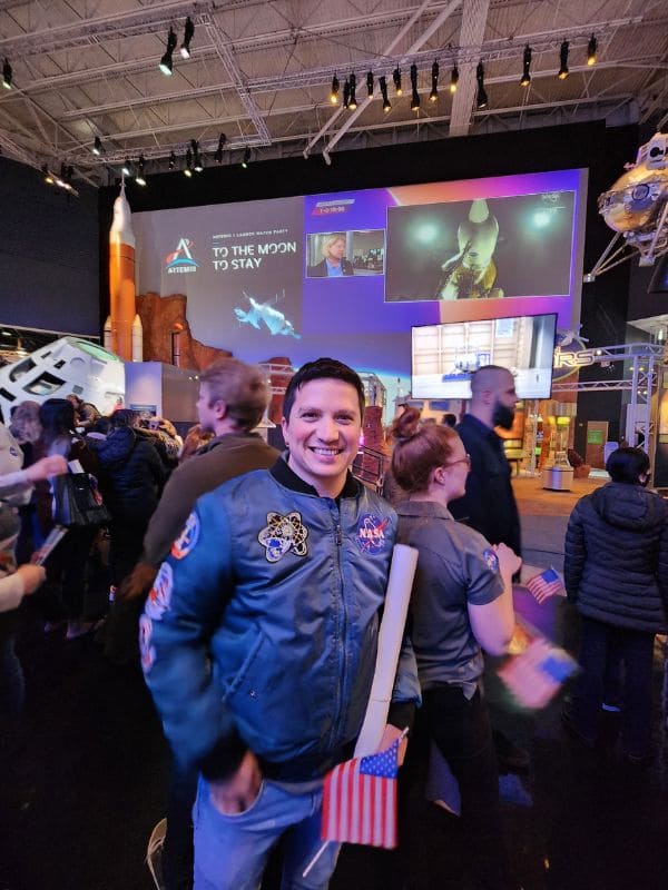 Manuel in a NASA jacket holds an American flag at an indoor space exhibit. Screens display space imagery with “To the Moon to Stay.” Crowded, lively atmosphere.