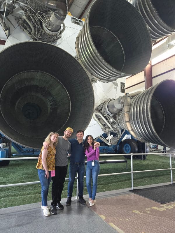 Four people stand closely together, smiling, beneath large rocket engines. The atmosphere is casual and joyful.