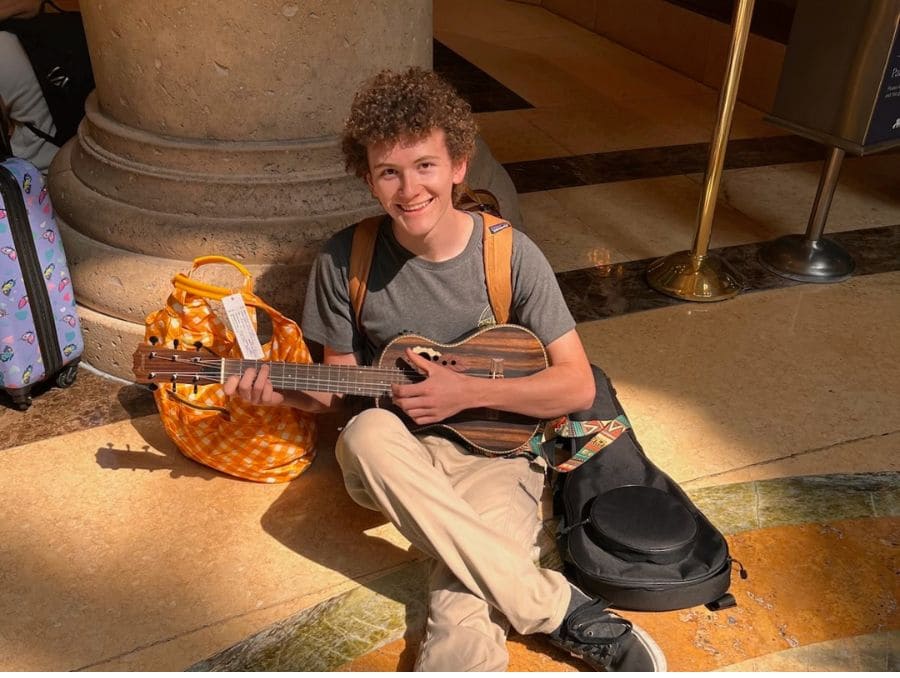A young person with curly hair smiles while sitting on a polished floor, playing a ukelele. They are by a large column, next to a colorful bag. 