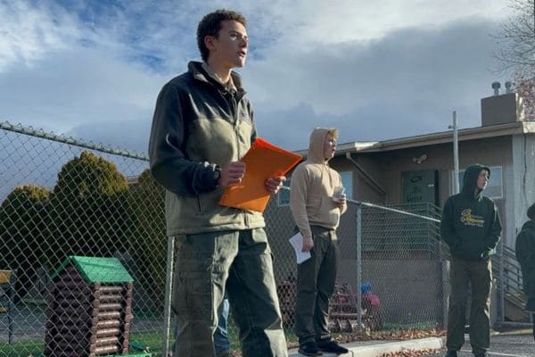 Young person outdoors, holding an orange folder, speaks confidently. Two others in the background listen. They stand beside a chain-link fence under a cloudy sky. 