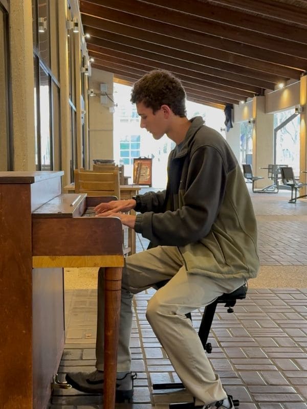 Young man in a green jacket plays a wooden piano in a covered outdoor walkway. The atmosphere is calm and relaxed with soft, natural lighting. 