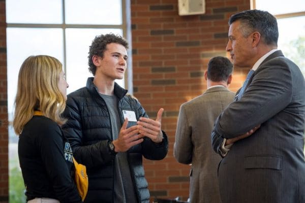 A young man gestures while speaking to an older man in a suit, with a woman listening closely beside them. The setting is casual yet professional, with a brick wall and large window in the background. 