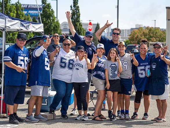 A group of enthusiastic University of Nevada, Reno fans pose together at a tailgate party in a parking lot, wearing Wolf Pack gear and jerseys. They are smiling, making hand signs, and standing in front of tents and vehicles decorated for the event, enjoying a sunny game day atmosphere.