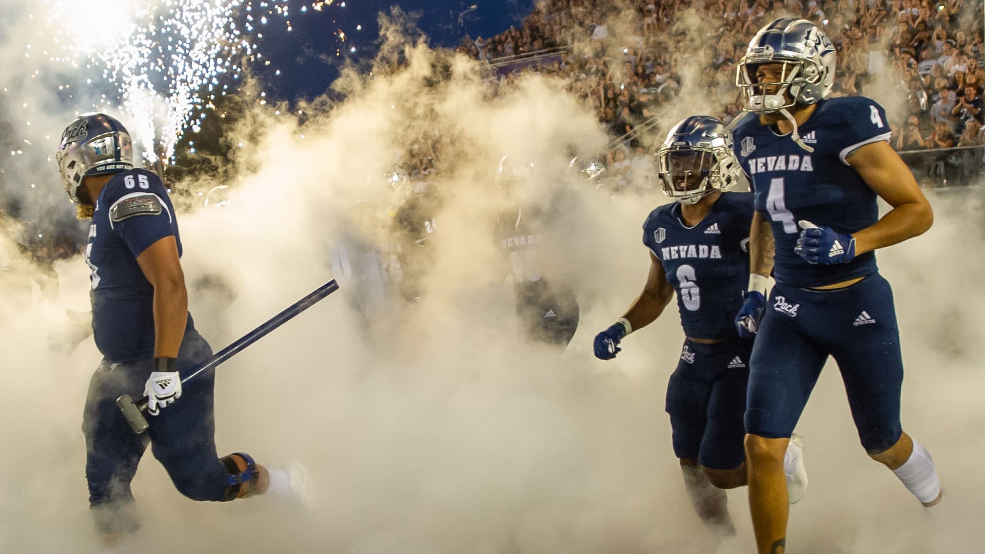A group of Nevada football players wearing blue jerseys and helmets run through smoke onto the field at Mackay Stadium.