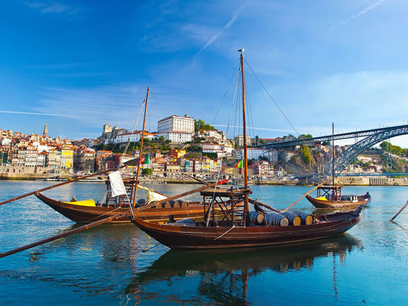 Two brown boats on a river heading under a bridge in front of a colorful town on a hill.