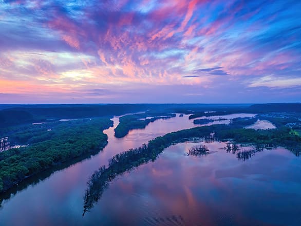 A glassy river filled with green trees under a blue and orange sunset sky.