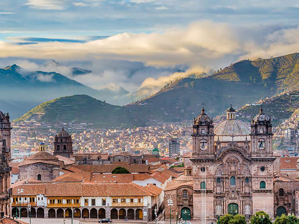 A large town with many small brown and red buildings and a cathedral in front of a large green mountain range covered by clouds.