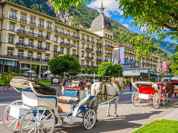 A long detailed yellow building on a town street with carriages in front of a green mountain range.
