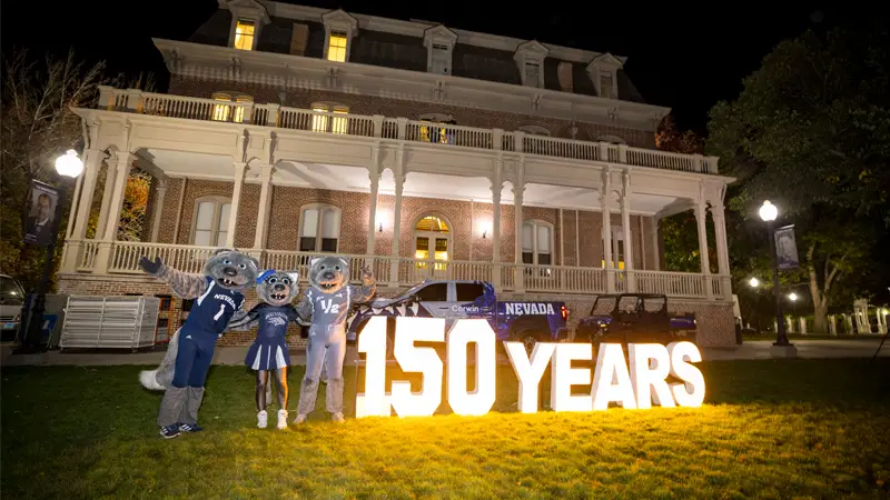 The three Nevada mascots, Alphie, Wolfie, Jr., and Luna stand next to a large lit sign that says "150 Years" in front of Morill Hall. A Nevada Corwin Ford truck is parked next to the building.