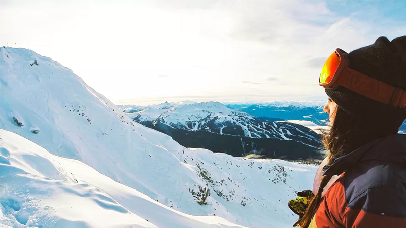 A woman wearing snow gear stares into the distance on a snowcapped mountain.