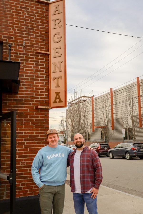 Two men stand smiling in front of a brick building with a vertical sign reading "ARGENTA." One man wears a light blue sweatshirt with the word "BUNKER" and a cap, while the other wears a plaid shirt over a white t-shirt. They stand on a city sidewalk with parked cars and buildings in the background.