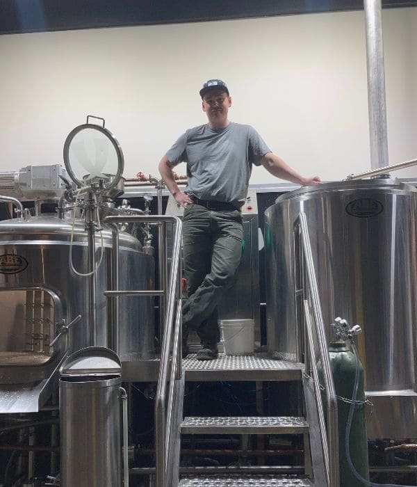 A man wearing a gray t-shirt, dark pants, and a baseball cap stands on a metal platform between large stainless-steel brewing tanks in a brewery. He has one hand on his hip and the other resting on a tank, looking confidently at the camera.
