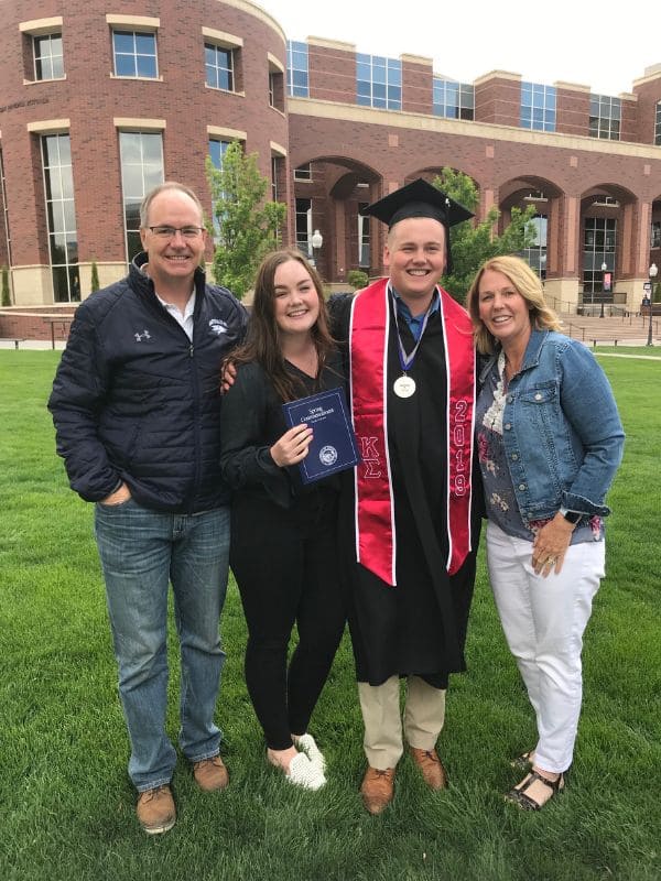 : A young man in a black graduation gown and cap, wearing a red and black stole, stands with his family on a green lawn in front of a university building. He holds a diploma while smiling alongside his parents and his sister.