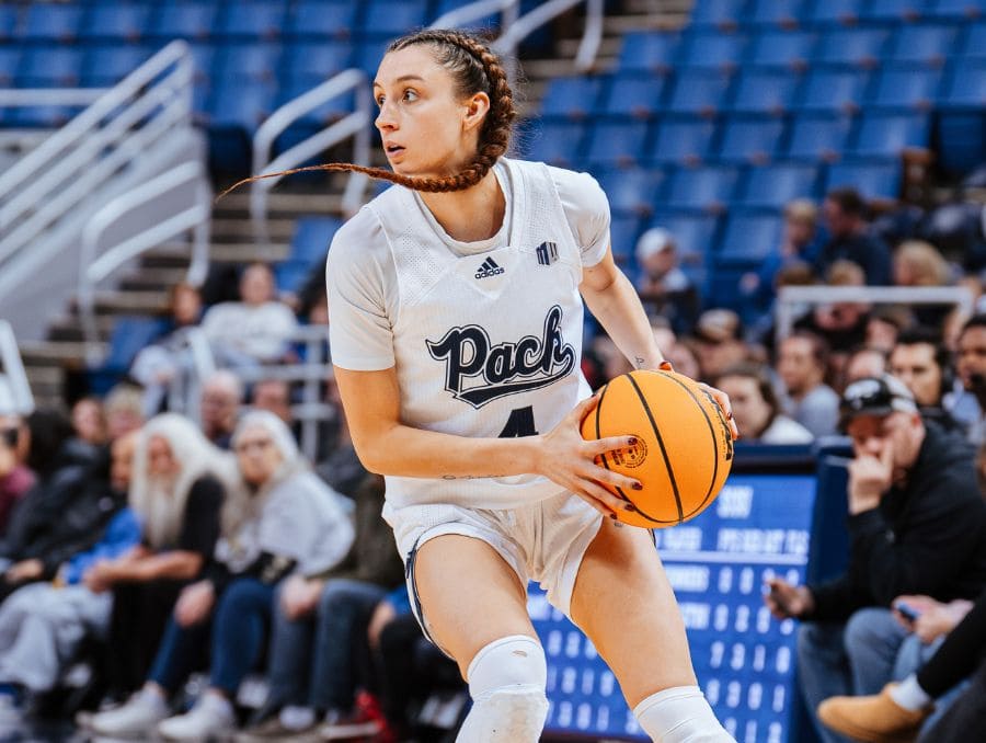A female basketball player with a braided ponytail, wearing a white "Pack" uniform, dribbles an orange basketball on a court.