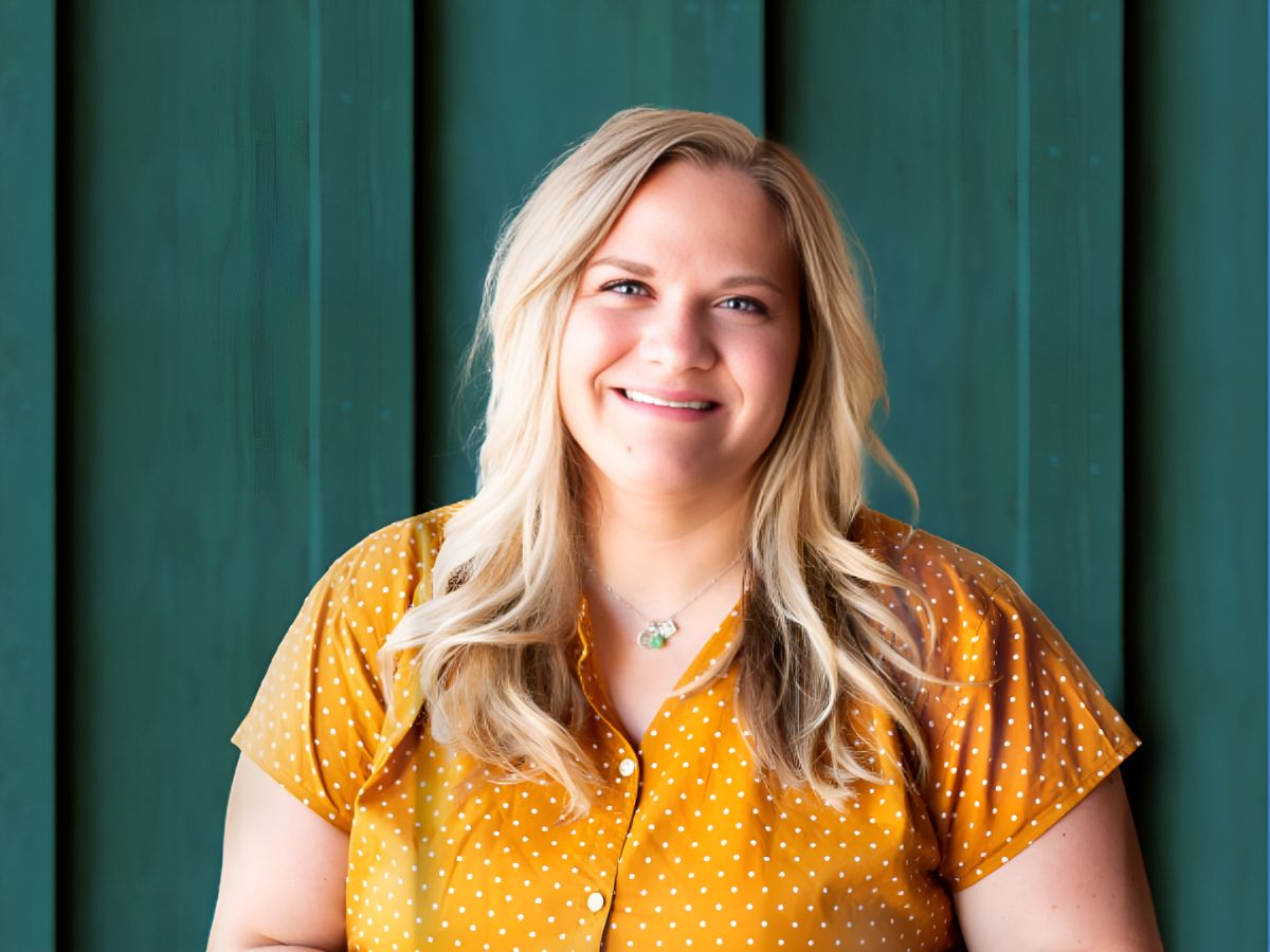 Andrea Thompson smiles and wears a yellow shirt with white polka dots while posing for a picture against a wooden background.