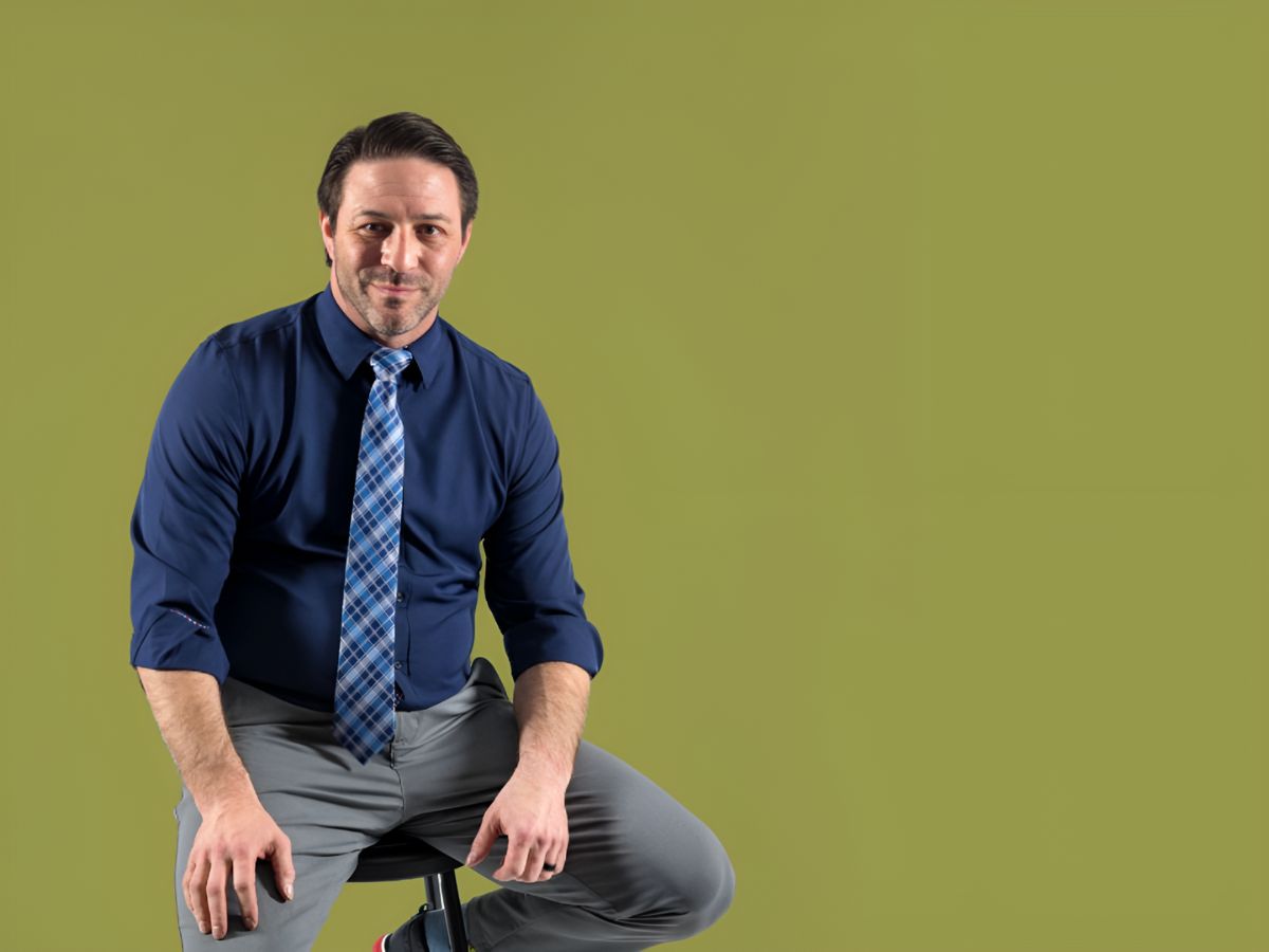 Joshua Fitzgerald, seated, wears a blue dress shirt, plaid blue tie, and smiles as he poses for a picture against a green background.
