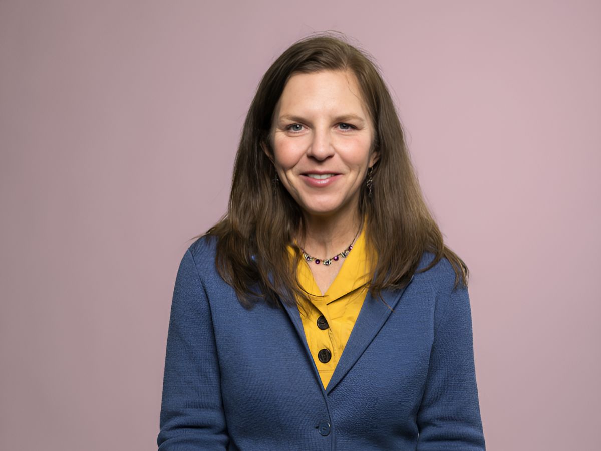 Marta Elliott wears a blue blazer, yellow shirt and necklace as she sits for a photo against a pink backdrop.