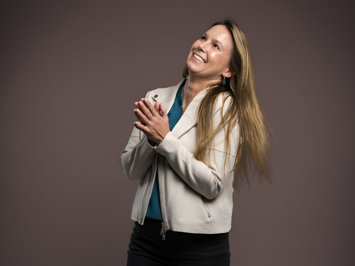 Shannon Burleson claps her hands together while wearing a gray blazer and blue shirt and smiles for a photograph while standing.