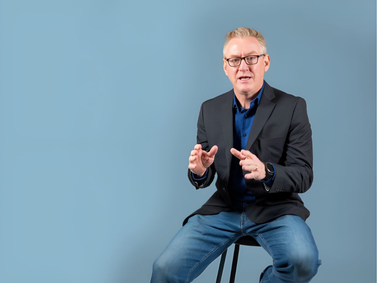 Steven Nicholas, seated, wears a black blazer, blue collared shirt, jeans, and glasses.