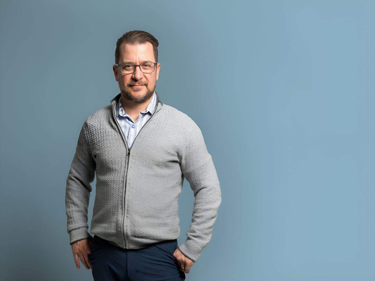 Yani Dickens wears a gray cardigan with a zipper, a white collared shirt, and glasses while posing for a picture against a backdrop with a blue gradient backround.