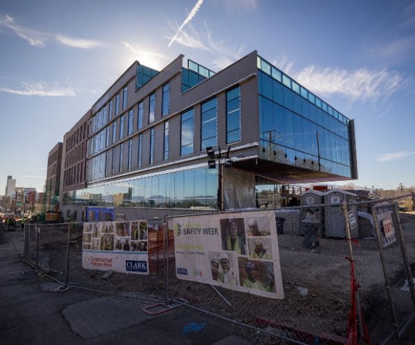 Construction site of a modern, glass-walled building under a clear blue sky. Safety banners and fences surround the area.
