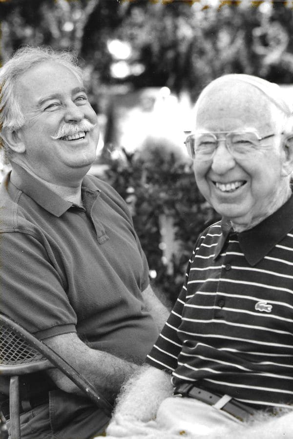 Two men, sitting outside in summer attire, smiling warmly. One wears a striped polo, the other a plain one.