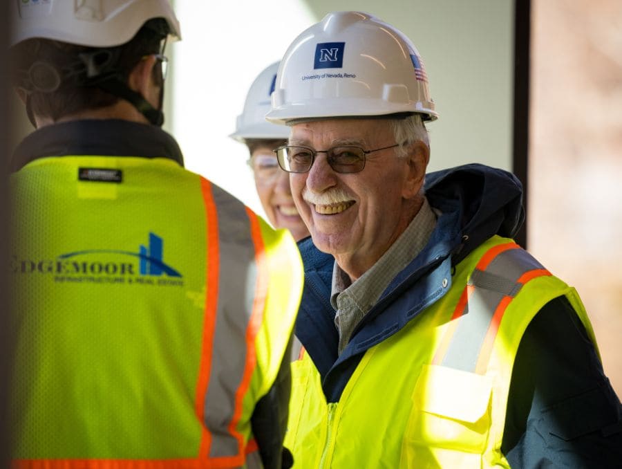 A smiling man in a hard hat and neon safety vest engages with colleagues at a construction site. 