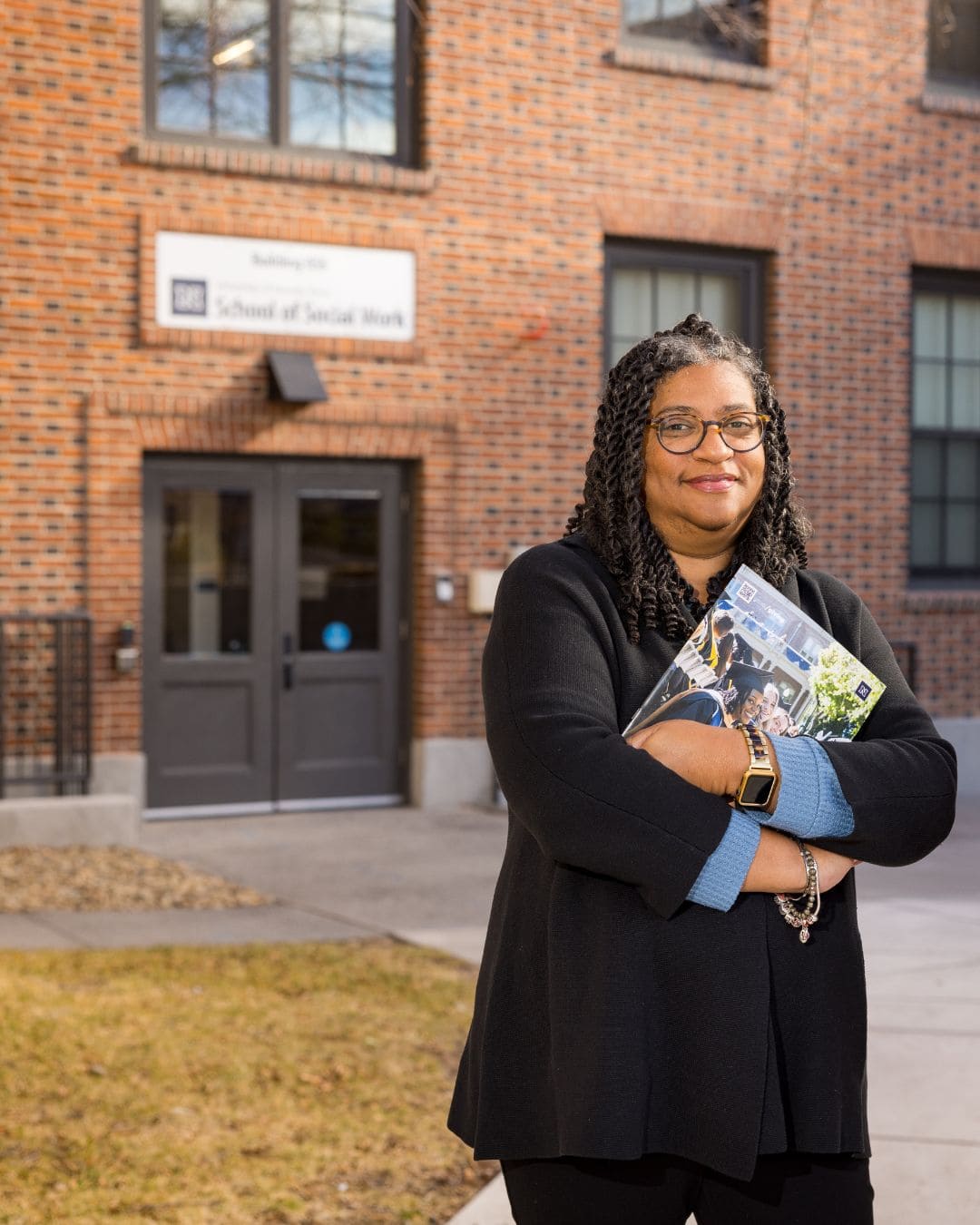 A woman wearing glasses holds a book while standing in front of a large building.