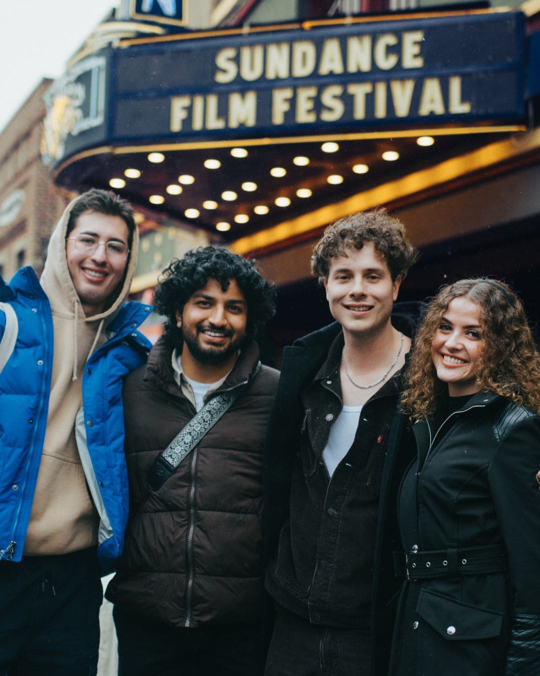 Four young people smile and pose in front of the Sundance Film Festival sign, showcasing their excitement for the event.