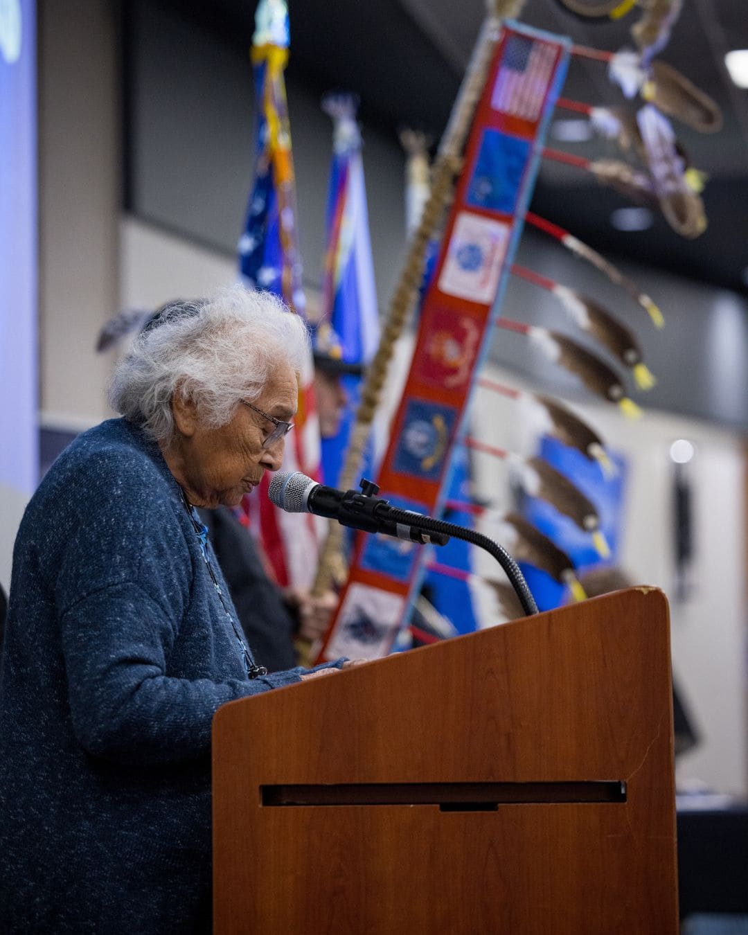 An elderly woman addresses an audience from a podium, flanked by various flags in the background.