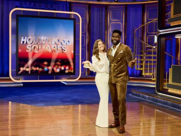Drew Barrymore and Nate Burleson pose together on a TV set. The “Hollywood Squares” logo is displayed on a screen beside them.
