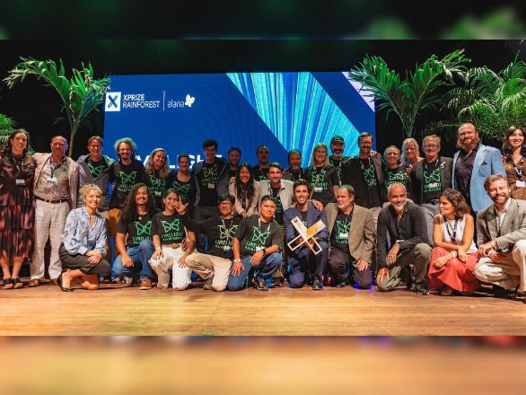 A group of people pose happily on stage with greenery in the background and a large screen displaying “XPRIZE Rainforest.”